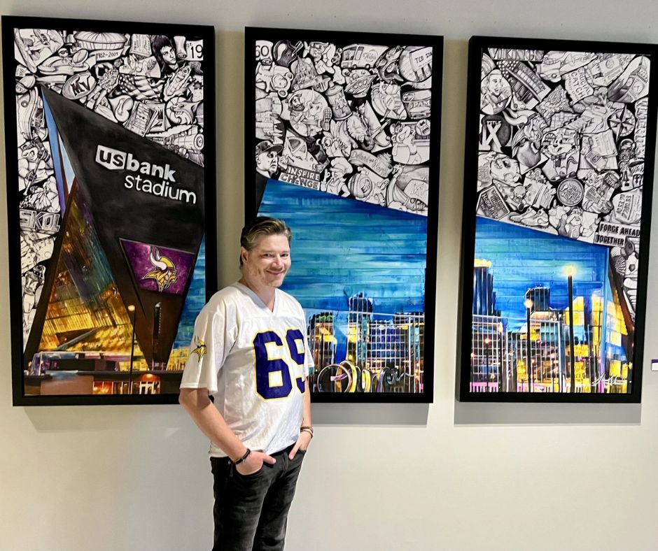 A middle-aged man with light brown hair, wearing a white Minnesota Vikings jersey with the number 69, stands in front of three vertically oriented pieces of framed artwork. The artwork features mixed media, with the bottom halves showing colorful, stylized scenes of Minneapolis or US Bank Stadium, and the top halves filled with black-and-white collage illustrations of Vikings players, fans, and pop culture references. The man is smiling and has his hands in his dark pants pockets